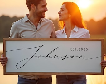 May include: A couple holds a white wooden sign with the text "Johnson EST. 2025" in black cursive script. The sign is framed in a light brown wood frame.