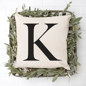 May include: A white pillow with a black letter K printed on it. The pillow is sitting in a woven basket with green leaves and white berries.