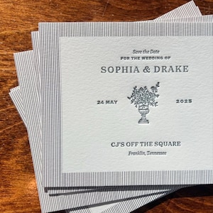 Pode incluir: Uma pilha de convites de casamento quadrados. Cada convite apresenta o texto "Save the Date" e os nomes "SOPHIA & DRAKE". A data "24 MAIO 2025" também está impressa. Os convites têm um design floral e o nome do local "CJ'S OFF THE SQUARE".