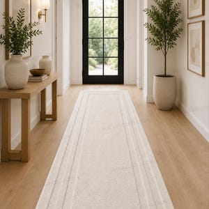 May include: A long, rectangular runner rug in a solid cream colour featuring a subtle, raised border design with concentric rectangular lines. The rug is positioned centrally on a light-toned wooden floor in a hallway leading to a black door.