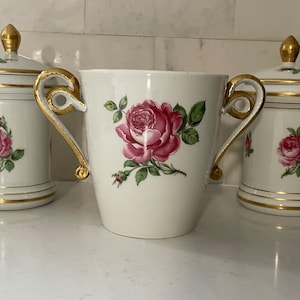 May include: A trio of white ceramic containers with gold detailing and rose motifs. The central piece is a handled vase, with two lidded canisters on either side. The roses are pink and red with green foliage.