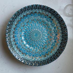 May include: A decorative, round plate with a turquoise floral design in the center. The plate has a silver and black ornate border. The plate is made of glass and is suitable for serving food or as a decorative piece.