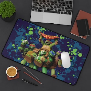 Puede incluir: Alfombrilla de ratón rectangular con las Tortugas Ninja. El diseño muestra a las cuatro tortugas sosteniendo una pizza, sobre un fondo azul oscuro con salpicaduras de pintura verdes y azules. También se ve un ratón blanco y un portátil.