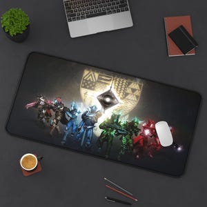 May include: A black rectangular gaming mousepad featuring a Destiny 2 design. The design includes a shield emblem and several characters in various colors. A white mouse is on the right side of the pad.