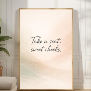 Puede incluir: Una impresión artística enmarcada con el texto "Take a seat, sweet cheeks," en una elegante escritura. El fondo presenta suaves lavados de acuarela en melocotón, verde y crema. La impresión se exhibe en una habitación con una paleta de colores neutros, una planta en maceta y un otomán beige.