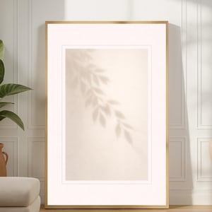 May include: A framed art print featuring a beige background with a leaf shadow. The print is in a gold frame and is leaning against a white wall. A large potted plant and a beige ottoman are in the foreground.