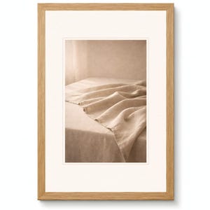 Puede incluir: Fotografía en tonos sepia enmarcada que muestra una tela de lino beige sobre una mesa. El lino tiene pliegues suaves. El marco es de madera marrón claro con un passe-partout blanco.