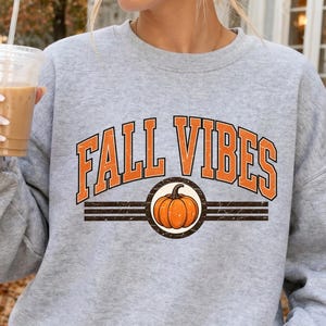 Pode incluir: Uma camisola cinzenta clara de gola redonda com o texto laranja FALL VIBES numa fonte universitária, centrado sobre um gráfico de uma abóbora laranja dentro de uma moldura circular com linhas pretas horizontais nos lados.