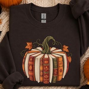 May include: A dark brown crewneck sweatshirt featuring a graphic of a pumpkin constructed from stacked vintage books with colorful spines, decorated with orange and green autumn leaves and a green stem, with a Gildan brand label at the neck.