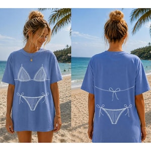 May include: A periwinkle blue short-sleeve t-shirt featuring a white line-art graphic of a bikini on the front and the back. The front displays the bikini top and bottom, while the back shows the bikini bottom and the tie strings of the top.
