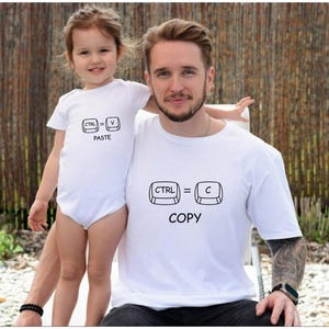 Op de afbeelding: Een vader en kind dragen bijpassende witte t-shirts. Het shirt van de vader zegt "CTRL = C COPY" en de romper van het kind zegt "CTRL = V PASTE". De shirts hebben zwarte grafische ontwerpen.
