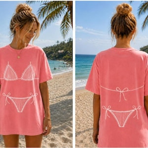 May include: A coral pink short-sleeve t-shirt featuring a white line art graphic of a bikini top on the front and a bikini bottom with a tie-side detail on the back. The shirt has a crew neckline and a relaxed, loose-fitting silhouette.