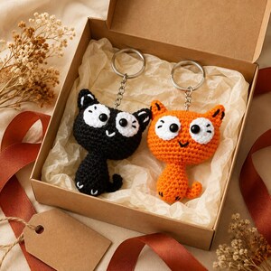 May include: Two crochet cat keychains, one black and one orange, each featuring large white eyes with black pupils and a small stitched mouth. The keychains are attached to silver metal rings and chains, presented inside a brown cardboard gift box.