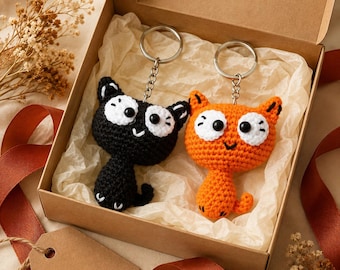 May include: Two crochet cat keychains, one black and one orange, each featuring large white eyes with black pupils and a small stitched mouth. The keychains are attached to silver metal rings and chains, presented inside a brown cardboard gift box.