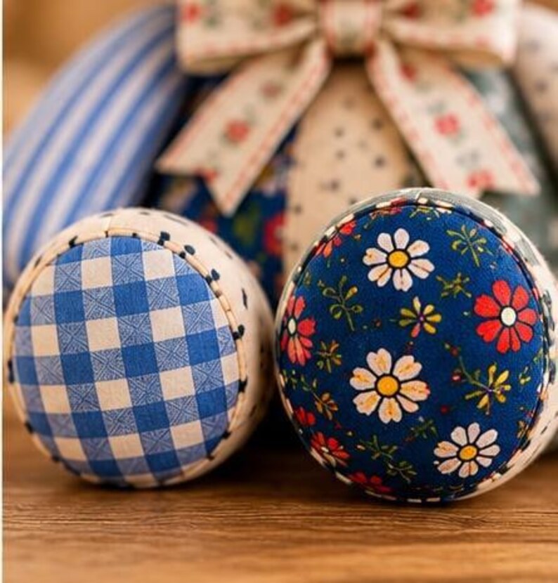 May include: Two round fabric pincushions featuring distinct patterns. One displays a blue and white gingham check, while the other showcases a dark blue background with a floral print of white daisies and red flowers. A fabric bow sits above.