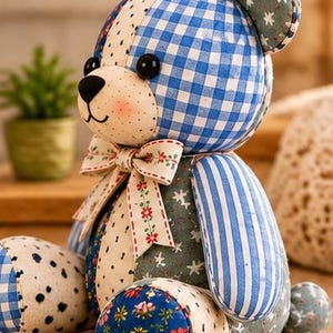 May include: A patchwork teddy bear crafted from various fabric patterns including blue and white gingham, blue stripes, floral prints, and star motifs. The bear features black button eyes, a black stitched nose, and a cream-coloured bow with red flowers.