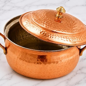 May include: A copper cooking pot with a lid, featuring a hammered texture. The pot has two handles and a decorative knob on the lid. The pot is open, revealing the interior. The copper pot is on a marble surface.