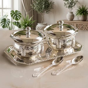 May include: A silver serving set comprising two lidded bowls, a rectangular tray, and two spoons. The bowls feature a hammered texture and tassel details. The spoons and tray have floral patterns.
