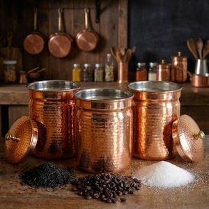 May include: Three copper storage jars with lids, next to piles of coffee beans, sugar, and tea leaves. The jars have a hammered texture and silver interiors. Copper pots and utensils are in the background, suggesting a kitchen setting.
