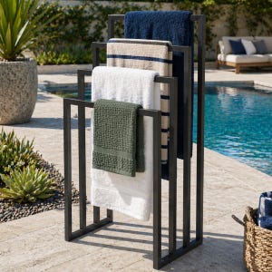 May include: A black metal towel rack holding several towels of different colors and textures. The towels include a dark blue, a beige striped, a white, and a green waffle-weave towel. The rack is outdoors near a pool.