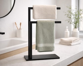 May include: A black metal towel rack with two levels, holding a beige and a sage green towel. The towels have a textured appearance and are folded neatly. The rack is on a white countertop in a bathroom setting.