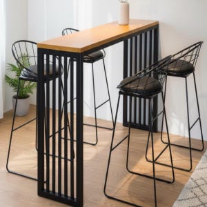 Puede incluir: Una mesa de bar moderna con una tapa de madera clara y un marco de metal negro, combinada con tres taburetes de bar negros con estructura de alambre y asientos acolchados negros. Un pequeño jarrón está sobre la mesa. La mesa mide unos 100 cm de largo.