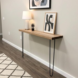May include: A light-colored wooden console table with black hairpin legs. The table is against a light gray wall and has a lamp, artwork, and a decorative object on top. The table is approximately 60 inches long.