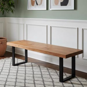 May include: A wooden bench with a light brown finish and black metal legs. The bench is placed on a patterned rug. The background features white wall panelling and abstract art. The bench's length is approximately 150 cm.
