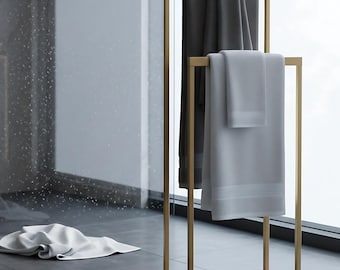 May include: A gold-colored metal towel rack holding two folded towels, one dark gray and one light gray. A white towel lies on the gray tiled floor. The scene is set in a modern bathroom with a large window.