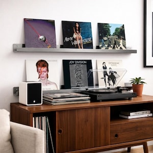 Könnte beinhalten: Ein modernes Schallplattenspieler- und Vinyl-Schrank-Set. Der Schrank ist aus dunklem Holz gefertigt und hat ein Regal mit mehreren ausgestellten Schallplatten. Ein weißer Lautsprecher steht auf dem Schrank.