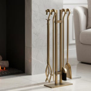 May include: A gold-coloured fireplace tool set with a stand. The set includes a poker, a brush, a shovel, and tongs. The tools are arranged vertically on a rectangular base. A marble fireplace and a white armchair are in the background.