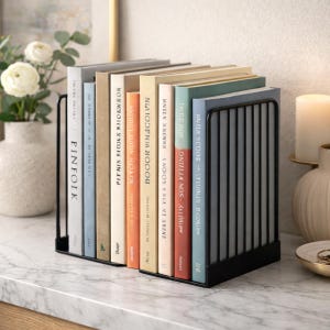 Puede incluir: Un organizador de libros de metal negro que contiene una colección de libros con lomos de varios colores. Los libros están dispuestos verticalmente, con los títulos visibles. El organizador se encuentra sobre una superficie de mármol, con un jarrón de flores y una vela en el fondo.