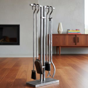 May include: A silver metal fireplace tool set with a stand. The set includes a poker, shovel, brush, and tongs. The tools have a simple, functional design and are arranged on a rectangular base. The background shows a fireplace and wooden furniture.