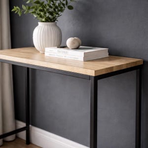 Puede incluir: Una consola moderna con una tapa de madera clara y una estructura de metal negro. Un jarrón blanco con vegetación, una pila de libros y una piedra decorativa están sobre la mesa. Los libros tienen la palabra "TINTERUS" en el lomo.