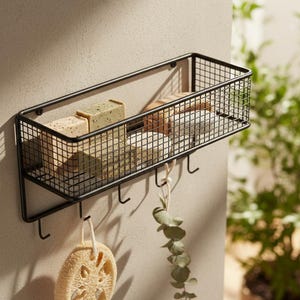 May include: Black metal wall-mounted storage basket with a rectangular design. The basket holds bars of soap and other bath accessories. Below the basket are hooks with a loofah and eucalyptus sprig hanging from them.