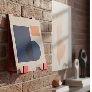 May include: A modern art print with geometric shapes in navy blue, orange, and beige, mounted on a red metal frame. The artwork is displayed against a brick wall, showcasing a contemporary design aesthetic. Other art pieces and decor are visible in the background.