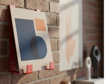 May include: A modern art print with geometric shapes in navy blue, orange, and beige, mounted on a red metal frame. The artwork is displayed against a brick wall, showcasing a contemporary design aesthetic. Other art pieces and decor are visible in the background.