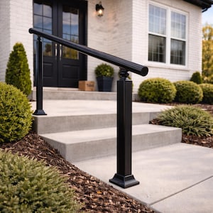 Op de afbeelding: Zwarte metalen leuning met een afgeronde bovenkant, geïnstalleerd op betonnen trappen. De leuning wordt ondersteund door vierkante palen met decoratieve accenten. De trap leidt naar een wit bakstenen huis met een zwarte deur.