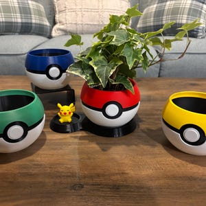 May include: Four spherical planters designed to resemble Poke Balls in blue, green, red, and yellow, each featuring a white center button and black trim. A small yellow Pikachu figure stands next to the red planter, which holds a green ivy plant.