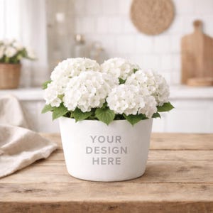 May include: A white ceramic pot with the text "YOUR DESIGN HERE" holds a lush arrangement of white hydrangea flowers with green leaves. The pot sits on a wooden table, with a kitchen setting in the background.