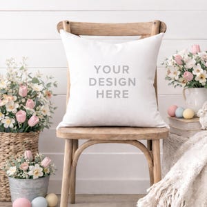 May include: A white square pillow with the text "YOUR DESIGN HERE" sits on a wooden chair. The scene includes floral arrangements in baskets and a pitcher, pastel Easter eggs, and a decorative blanket, creating a spring or Easter theme.