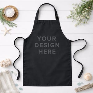 May include: A black apron with the text "YOUR DESIGN HERE" printed in gray. The apron has a neck strap and waist ties. The apron is displayed on a white wooden surface with decorative elements.