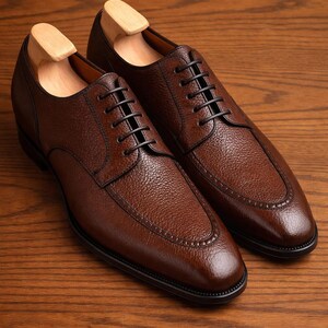Pode incluir: Um par de sapatos de couro castanho com atacadores pretos e um acabamento texturizado. Os sapatos têm um design clássico com biqueira arredondada e são exibidos numa superfície de madeira. São inseridas formas de sapato.