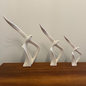 May include: Three white ceramic bird sculptures in a row, each with an abstract design. The sculptures are on a wooden surface. The largest sculpture is on the left, and the smallest is on the right. The background is a neutral wall.
