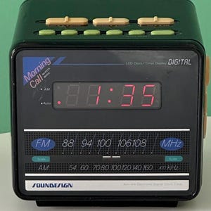 May include: A retro digital alarm clock in black and green. The red LED display shows the time. It has FM/AM radio, a buzzer, and the words "Morning Call". The clock measures approximately 10 cm in width.