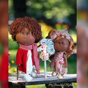 May include: Two Blythe dolls stand side-by-side. One doll has brown hair, a red coat, and a striped scarf. The other doll has blonde hair and a floral dress. They are holding a unicorn toy. The dolls are on a wooden surface.