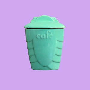 Può includere: Un barattolo in ceramica turchese con coperchio, con la scritta "cafe" in rilievo. Il barattolo ha un design unico a strati sui lati e una parte superiore decorativa. Lo sfondo è di colore lavanda.