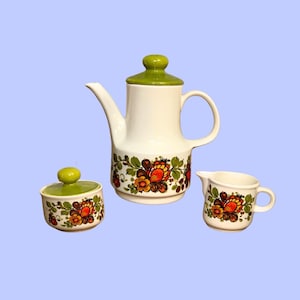 Puede incluir: Un juego de café de cerámica que consta de una cafetera alta con tapa verde, un azucarero con tapa verde a juego y una pequeña jarra de leche. Cada pieza es blanca y presenta una banda decorativa con motivos florales en naranja, amarillo y verde.