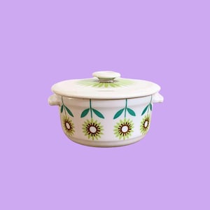 Puede incluir: Una fuente de cerámica blanca con tapa, adornada con un patrón repetido de diseños florales verdes y marrones. El plato tiene dos asas pequeñas y un patrón floral a juego en la tapa. El fondo es de color púrpura sólido.