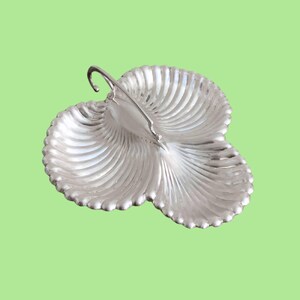 Könnte beinhalten: Silberfarbene Servierschale mit drei Fächern und einem Griff. Die Schale hat einen gewellten Rand und ein geripptes, fächerartiges Design. Der Griff ist nach oben gebogen. Die Schale befindet sich auf einem hellgrünen Hintergrund.